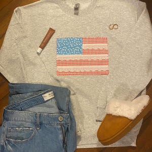 May include: A grey sweatshirt featuring a red, white and blue American flag design. The flag is made of fabric with a gingham pattern and lace trim. The sweatshirt is styled with blue jeans and brown slippers.