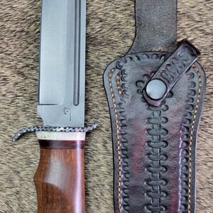 Peut inclure: Un couteau à lame fixe avec une lame en acier gris, un manche en bois avec des accents blancs et rouge foncé, et une garde en métal texturé. À côté se trouve un étui en cuir marron foncé avec des coutures décoratives et un bouton-pression.