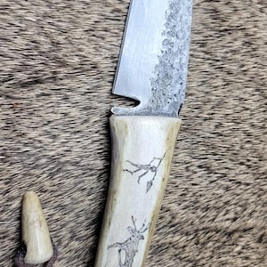 Peut inclure: Un couteau à lame fixe avec une lame en acier argenté à finition martelée présentant une petite encoche près de la garde. Le manche est en bois de cerf clair avec des dessins au trait noir gravés d'un cerf et d'une silhouette, avec une lanière.