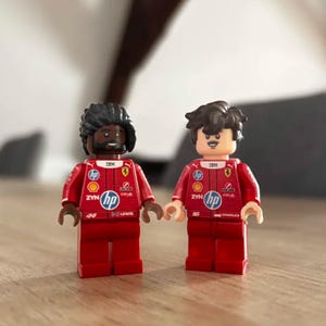 Pode incluir: Duas minifiguras de plástico vestindo trajes de corrida vermelhos com logotipos de patrocinadores, incluindo HP, IBM e Shell. A figura da esquerda tem pele escura, barba e cabelo preto, marcada com 44 e Lewis. A da direita tem pele clara e cabelo castanho, marcada com 16 e Charles.