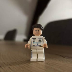 May include: A plastic minifigure with dark brown hair and a neutral expression, wearing a white racing suit. The suit features various printed logos including Barclays and Atlassian on the chest, with Atlassian also printed on the right leg.