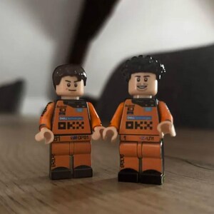 May include: Two plastic minifigures wearing orange racing suits with black accents. The suits feature printed logos including Dell Technologies and OKX. One figure has short brown hair, while the other has curly black hair. Both figures have articulated limbs.