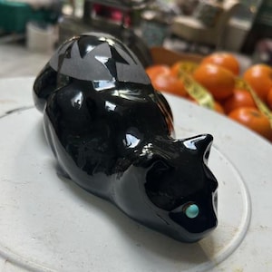 May include: A glossy black ceramic cat figurine with a turquoise eye. The cat has a geometric design on its back. The figurine is resting on a white surface.