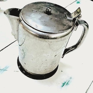 May include: A stainless steel pitcher with a lid and handle. The pitcher has a spout for pouring and sits on a black base. The lid has a small knob on top. The pitcher is designed for serving beverages.