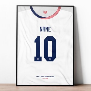 May include: A white framed print of a football shirt with a blue number 10 and the text "NAME" above the number. The shirt has a red, white and blue stripe pattern around the neck and the text "THE STARS AND STRIPES Since 1913" at the bottom.