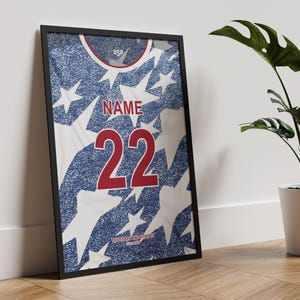 May include: Framed print of a USA soccer jersey design. The jersey features a blue and white star pattern with the number 22 in red. The word "NAME" is above the number. The top of the jersey has the word "USA".