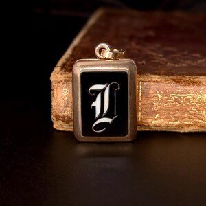Peut inclure: Pendentif rectangulaire en or avec un centre en émail noir orné d'un « I » blanc stylisé. Le pendentif est doté d'une petite boucle en haut pour une chaîne. L'arrière-plan est une image floue d'un livre.
