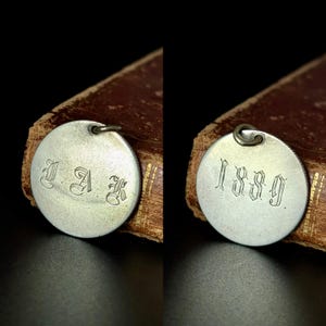 Può includere: Due ciondoli circolari in metallo argentato. Un ciondolo ha le iniziali "I A K" in un carattere ornato. L'altro ciondolo ha l'anno "1889" in un carattere simile. Entrambi i ciondoli hanno un piccolo anello nella parte superiore.