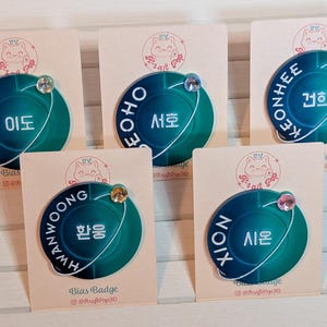 Könnte beinhalten: Fünf runde, blaue und grüne Emaille-Pins mit weißer Beschriftung. Jeder Pin zeigt ein anderes Mitglied einer K-Pop-Gruppe. Die Pins werden auf weißem Karton mit einer rosa-weißen Katzenillustration und dem Text "Bias Badge" angezeigt.