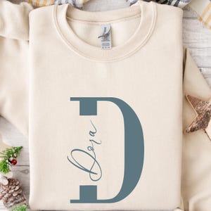May include: Cream-colored sweatshirt with a large teal letter "D" and the name "Dora" in a script font. The sweatshirt is made of a soft material and is likely for casual wear. The design is simple and elegant.
