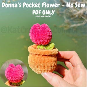 May include: A handmade crochet pocket flower in a small orange pot. The flower is a vibrant pink, with green leaves. The text "Donna's Pocket Flower - No Sew PDF ONLY" is at the top of the image.