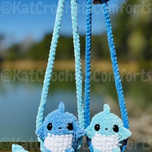 Pu&ograve; includere: Due borsette a forma di squalo all'uncinetto blu con occhi neri e pance bianche sono sospese da cinghie blu. Le borsette sono tenute contro uno sfondo sfocato di acqua e alberi. Il testo "@KatCrochetComforts" &egrave; visibile nell'immagine.