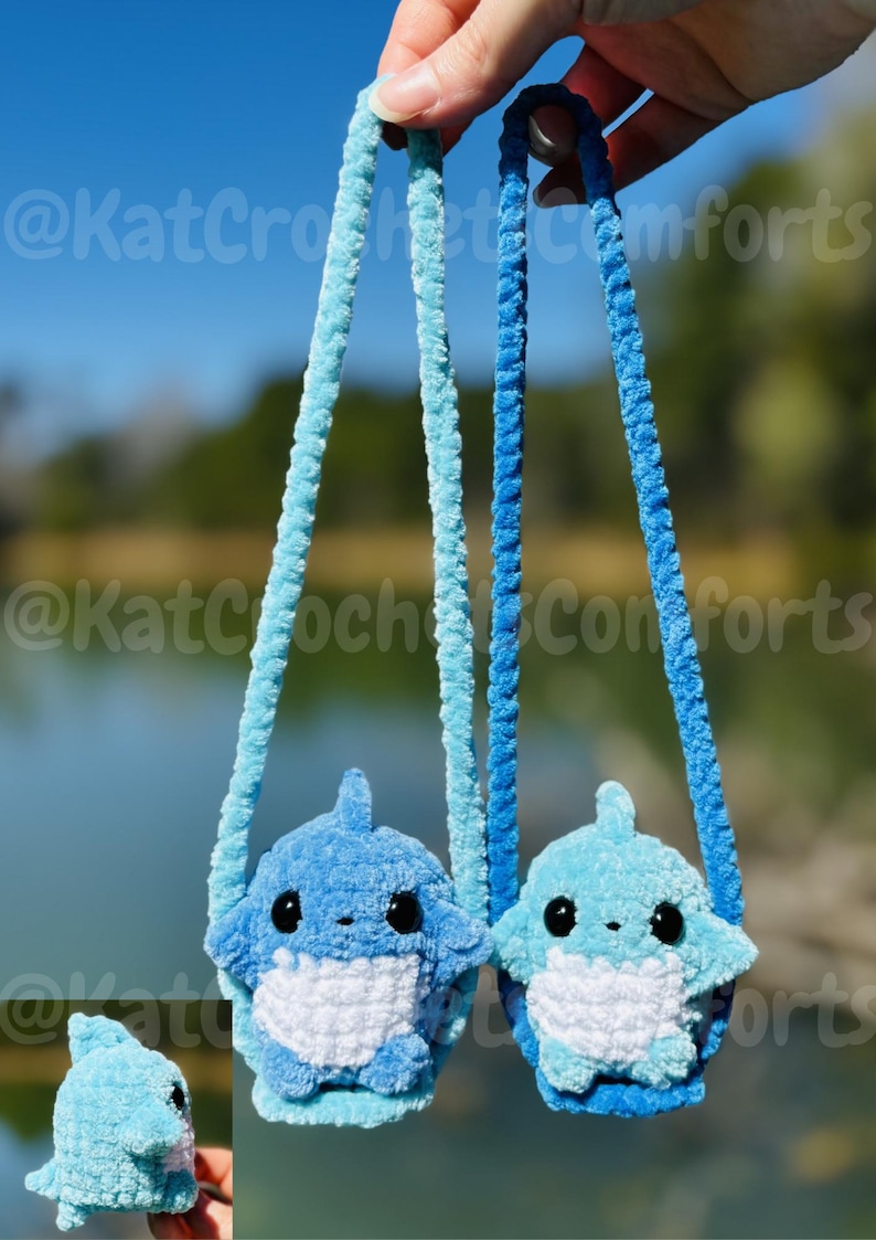 Pu&ograve; includere: Due borsette a forma di squalo all'uncinetto blu con occhi neri e pance bianche sono sospese da cinghie blu. Le borsette sono tenute contro uno sfondo sfocato di acqua e alberi. Il testo "@KatCrochetComforts" &egrave; visibile nell'immagine.