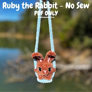Op de afbeelding: Een handgemaakt, bruin pluche konijnengezicht met roze binnenoren en zwarte ogen, hangend aan een lichtblauw koord. De tekst "Ruby the Rabbit - No Sew PDF ONLY" is zichtbaar. Het is een haakproject.