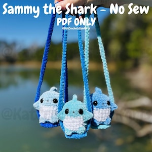 May include: Three crocheted shark plushies in varying shades of blue, each with black eyes and white bellies, suspended by blue cords. The text "Sammy the Shark - No Sew PDF ONLY" is at the top, with the seller's name below.