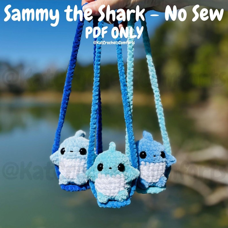 Pu&ograve; includere: Tre peluche a forma di squalo all'uncinetto in varie tonalit&agrave; di blu, ciascuno con occhi neri e pance bianche, sospesi da cordoncini blu. Il testo "Sammy the Shark - No Sew PDF ONLY" &egrave; in alto, con il nome del venditore sotto.