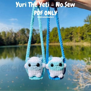 May include: Two small, white and blue crocheted Yeti plushies with black eyes and blue straps. The text "Yuri The Yeti - No Sew PDF ONLY" is at the top of the image. The plushies are hanging in front of a lake.