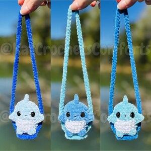 Pu&ograve; includere: Tre borse a forma di squalo in peluche in varie tonalit&agrave; di blu. Ogni borsa ha un cinturino blu abbinato. Le borse sono tenute contro uno sfondo sfocato verde e blu. Il testo "@ka CrochetsComforts" &egrave; visibile.