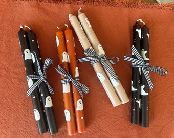 Puede incluir: Conjuntos de velas cónicas de Halloween en negro, naranja y beige. Cada vela está decorada con diseños de fantasmas blancos y atada con una cinta a cuadros en blanco y negro. Las velas están dispuestas sobre una superficie naranja.