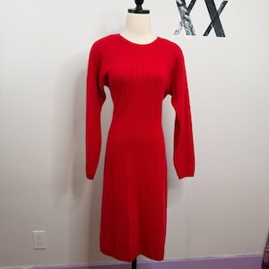 May include: A vibrant red long-sleeve sweater dress featuring a classic cable knit pattern throughout the garment. The dress has a round neckline and a straight silhouette that falls to a midi length, displayed on a mannequin form.