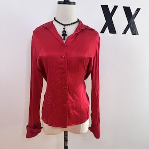 May include: A long-sleeved, red button-down shirt with a collar. The shirt is made of a shiny, silky material. A black beaded necklace with a pendant is worn with the shirt. The shirt is displayed on a mannequin.