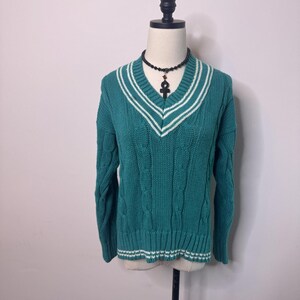 May include: A teal cable-knit sweater featuring a V-neckline with white striped trim. The long-sleeved garment has a ribbed hem and cuffs with white stripes. A black beaded necklace with an ankh pendant is displayed around the neck area.
