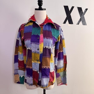 Può includere: Una camicia patchwork con colletto rosso e chiusura con bottoni. La camicia è realizzata con quadrati di tessuto di vari colori, tra cui viola, verde, giallo e marrone. La camicia è esposta su un manichino.