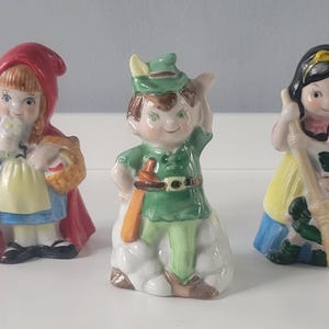 May include: Three ceramic figurines of storybook characters: Little Red Riding Hood, Peter Pan, and Snow White. The figurines are brightly colored and detailed, with Red Riding Hood holding a basket, Peter Pan with a sword, and Snow White with a broom.
