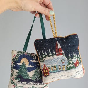 May include: Two square, fabric-covered ornaments featuring detailed needlepoint-style winter scenes. One depicts a horse-drawn sleigh in a snowy forest under a full moon, while the other shows a church and houses in a snowy village setting.