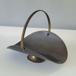 May include: A dark brown metal basket with a curved, open-top design and a single arched brass handle. The basket is supported by a circular brass pedestal base and features a small metal rivet on the side of the curved body.
