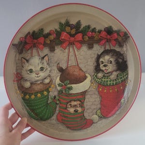 May include: Round metal tray with a festive Christmas design. The tray features a fireplace scene with three stockings, each holding a kitten, puppy, and hedgehog. The tray has a red rim and is decorated with bows and greenery.