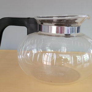 May include: A clear glass coffee carafe with a black plastic handle and a silver-toned metal band around the top rim. The carafe features the white text BUNN printed on the front, along with a small white graphic icon below the brand name.
