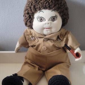 May include: A handmade doll dressed in a tan uniform with a matching hat and black shoes. The doll has a brown crocheted hairstyle and is holding a small pipe. The doll's face is made of fabric with embroidered features.