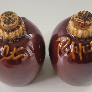 May include: A pair of brown ceramic salt and pepper shakers. Each shaker is round with a cork stopper and the words "Salt" and "Pepper" in a lighter brown script. The shakers have a glossy finish.