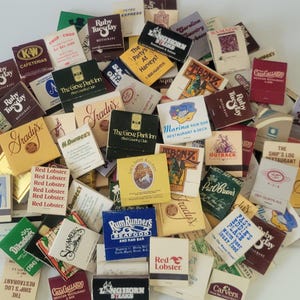 May include: A collection of vintage matchbooks from various restaurants and businesses. The matchbooks feature a variety of colors, logos, and text, including "Ruby Tuesday," "Red Lobster," and "Outback." A nostalgic collection.