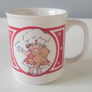 May include: White ceramic mug featuring a cartoon illustration of a girl in a chef's hat holding a teapot and a teddy bear. The mug has a red border with white floral accents and a curved handle.
