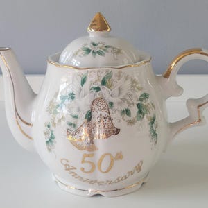 May include: White ceramic teapot with gold trim, a lid, and a handle. The teapot is decorated with a floral and dove design, and the words "50th Anniversary" are written in gold script. The spout and lid finial are also gold.