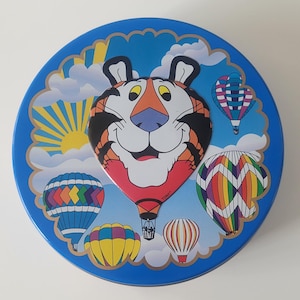 May include: A round, blue tin featuring a cartoon tiger face with a red and black hot air balloon body. The tin is decorated with colorful hot air balloons, clouds, and a sun against a blue sky background.