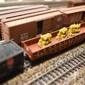 May include: A brown model train flatcar with the text P.E.N.N.S.Y.L.V.A.N.I.A. 363242 printed on the side, carrying two small yellow industrial engine models. The flatcar is positioned on model train tracks next to a brown boxcar labelled NYC 189000.