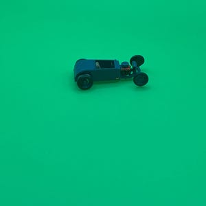 Puede incluir: Un pequeño coche de juguete azul oscuro con ruedas negras y un motor detallado. El vehículo en miniatura se encuentra sobre un fondo verde brillante, mostrando su diseño clásico de hot rod. El tamaño compacto y el color vibrante del coche lo hacen visualmente atractivo.