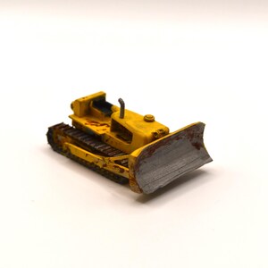 Peut inclure: Un petit bulldozer jouet jaune doté de chenilles marron foncé, d'une lame avant en métal argenté avec de la rouille visible et d'un tuyau d'échappement vertical. La carrosserie de l'engin de chantier présente des signes d'usure et d'écaillage.