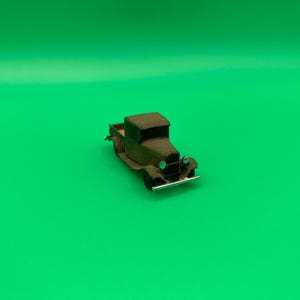 Könnte beinhalten: Ein kleiner olivgrüner Pickup-Truck mit schwarzen Rädern und einer silbernen Frontstoßstange. Der Spielzeug-Truck hat ein detailliertes Design, einschließlich Kabine, Pritsche und Scheinwerfern. Der Truck steht vor einem hellgrünen Hintergrund.