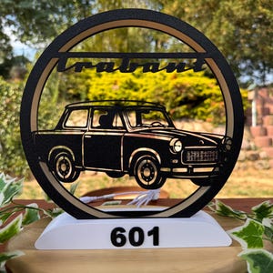 Könnte beinhalten: Ein kreisförmiges, schwarz-beiges Dekoschild mit der Silhouette eines Trabant-Autos und dem Schriftzug "trabant" oben. Das Schild ist auf einem weißen Sockel montiert, auf dessen Vorderseite die Zahl "601" in Schwarz zu sehen ist.