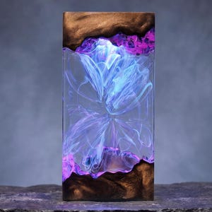 May include: A rectangular, illuminated resin art piece. The top and bottom sections are a dark wood grain, framing a clear resin center. Within the resin, a swirling, abstract design in shades of blue, purple, and white is visible.