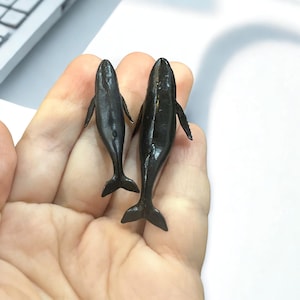 May include: Two small, black whale figurines with fins and tail flukes are held in a hand. The whales are positioned vertically, with their bodies facing upwards. The background is a white surface.