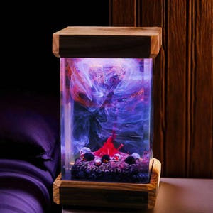 May include: A rectangular wooden-framed lamp with a clear, illuminated interior. The interior features a swirling, colorful design in shades of purple, red, and blue, with small skull accents at the base. The lamp is designed to sit on a flat surface.