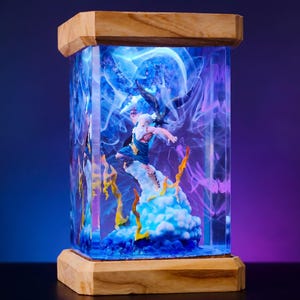May include: A rectangular resin lamp with a wooden base and top. Inside, a detailed figurine of a character in action is surrounded by blue and purple swirling patterns, with yellow and orange accents. The lamp emits a soft, ambient glow.