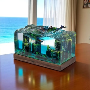 Puede incluir: Una escultura rectangular de resina que representa una escena submarina con ruinas arquitectónicas, coral y una ballena. La escultura está sobre una base de madera, con tonos azules y verdes. El fondo muestra una vista del océano.