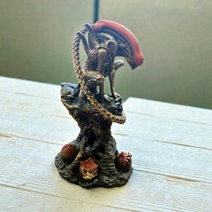 Könnte beinhalten: Detaillierte Figur einer Alien-Kreatur mit einem rot-orangen Kopf und einem segmentierten Schwanz. Die Figur steht auf einem strukturierten, braunen Sockel mit drei eiförmigen Strukturen. Die Farbpalette umfasst Braun-, Bronze- und Rottöne.