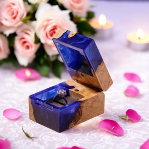 May include: A blue and wood ring box with a ring inside. The box is open and the ring is visible. The box is surrounded by rose petals.